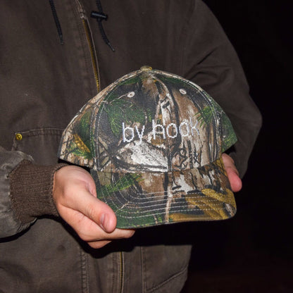 Camo print baseball hat with the words "by hook" being held by someone.