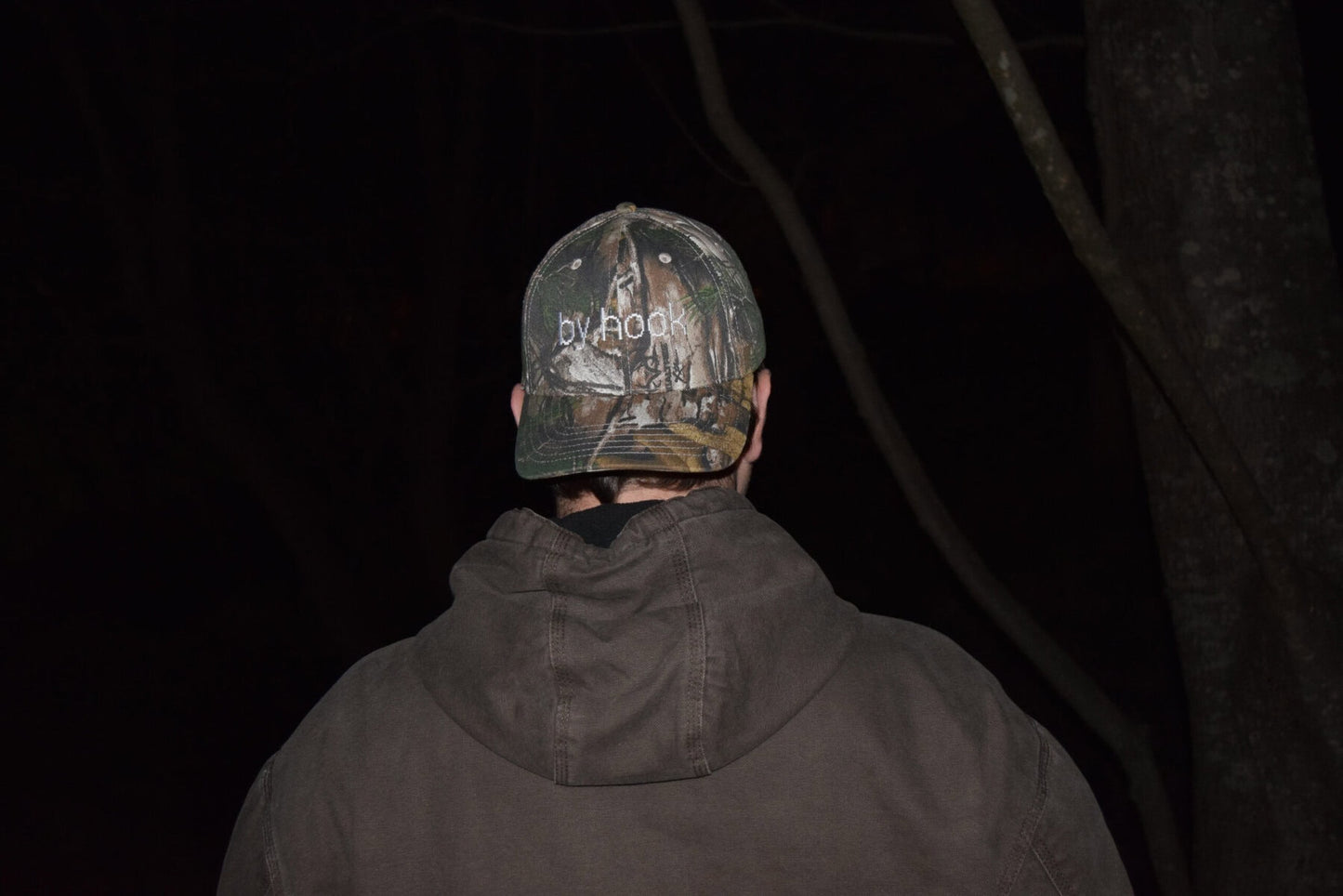 Camo print baseball hat with the words "by hook" being worn by someone.