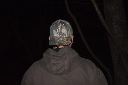 Camo print baseball hat with the words "by hook" being worn by someone.